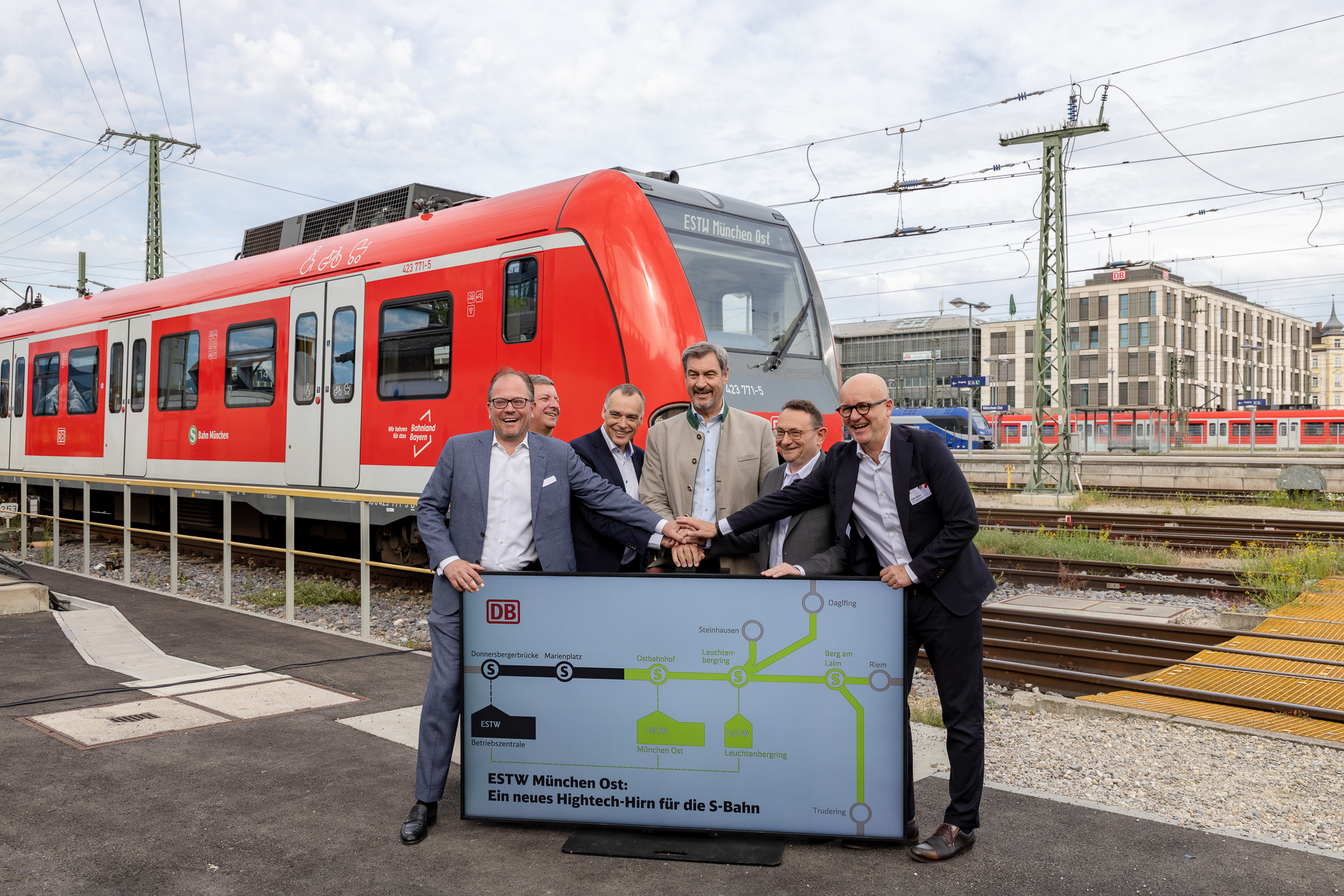 Bild zeigt sechs Männer, die gemeinsam einen Buzzer herunterdrücken, unterhalb des Buzzers befindet sich ein Bildschirm, auf dem ein Teil einer Strecke schwarz und ein Teil grün dargestellt ist. Im Hintergrund Gleise und eine S-Bahn